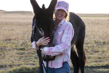 Woman in a pink shirt and hat hugging a black donkey in a field.