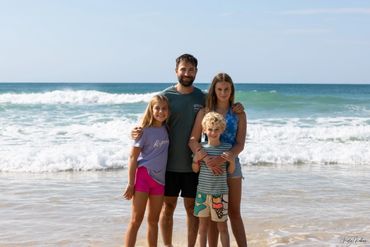 A family of four posing happily by the ocean shore on a sunny day.