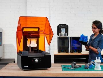 Operario trabajando con impresoras 3D y piezas recién impresas en una mesa de trabajo.