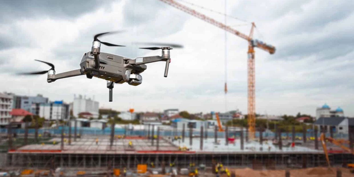 A drone flies over a construction site with a crane and buildings in the background.