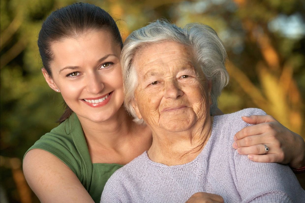 Young woman embraces elderly woman outdoors, both smiling warmly.
