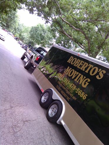 A truck towing a trailer for Roberto's Moving company parked on a tree-lined street.