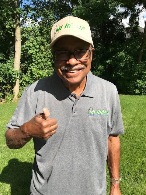 Smiling man giving thumbs up in MI-BAM branded shirt and cap outdoors.