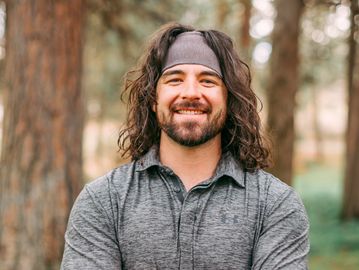 Smiling man with long hair and headband outdoors in a forest.