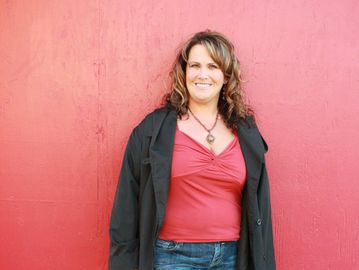 Woman in casual outfit stands against a red wall, smiling.
