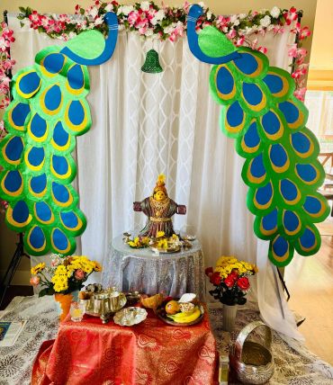 Colorful peacock decorations frame a festive altar with flowers and offerings.