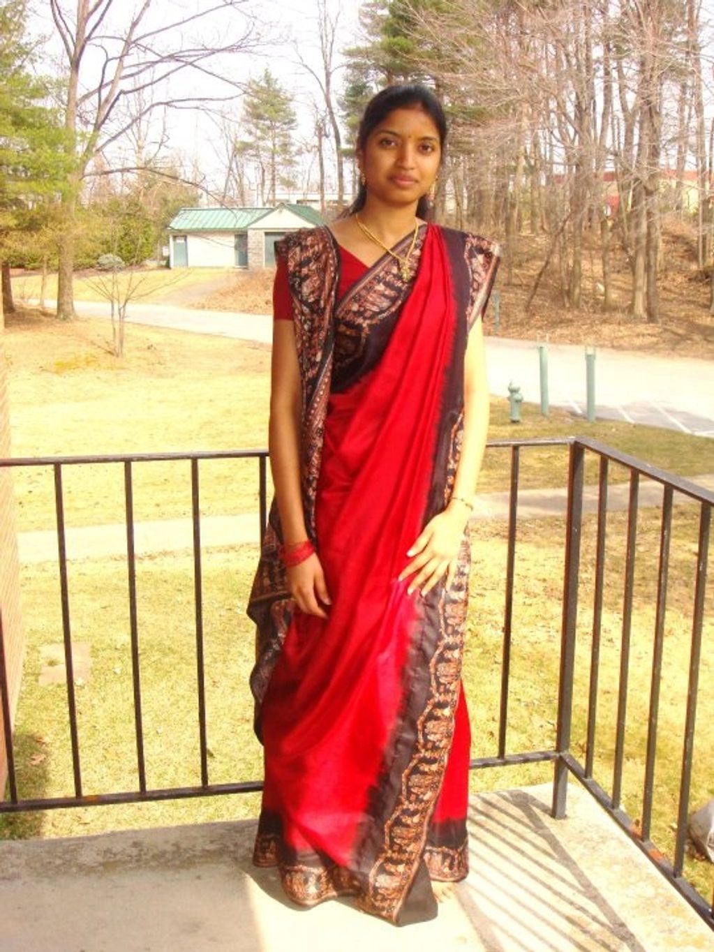 A woman in a red and black saree standing on a balcony in daylight.