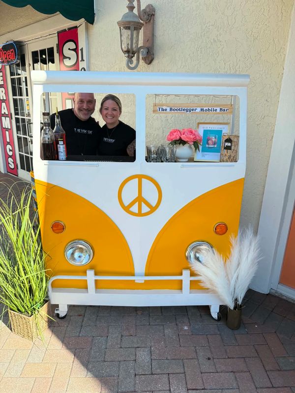Two people smiling behind a yellow and white mobile bar shaped like a vintage van.
