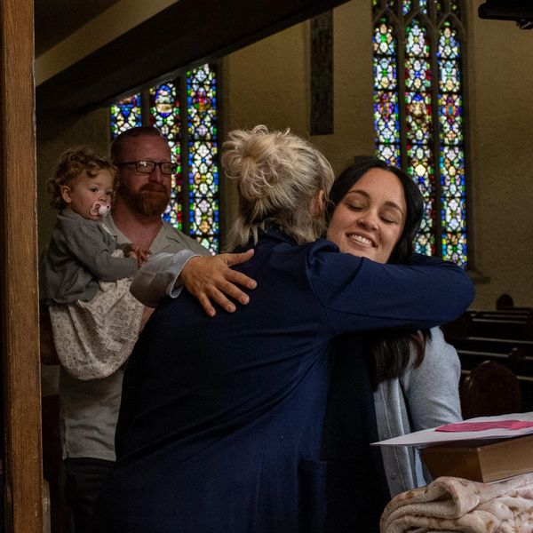Two women warmly embrace inside a church, with a man holding a child nearby.