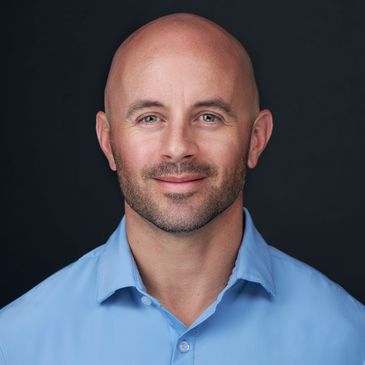 Smiling bald man in a blue shirt against a dark background.