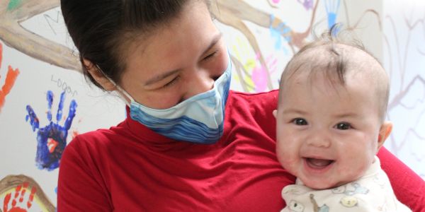 Woman in red with mask smiles at happy baby.