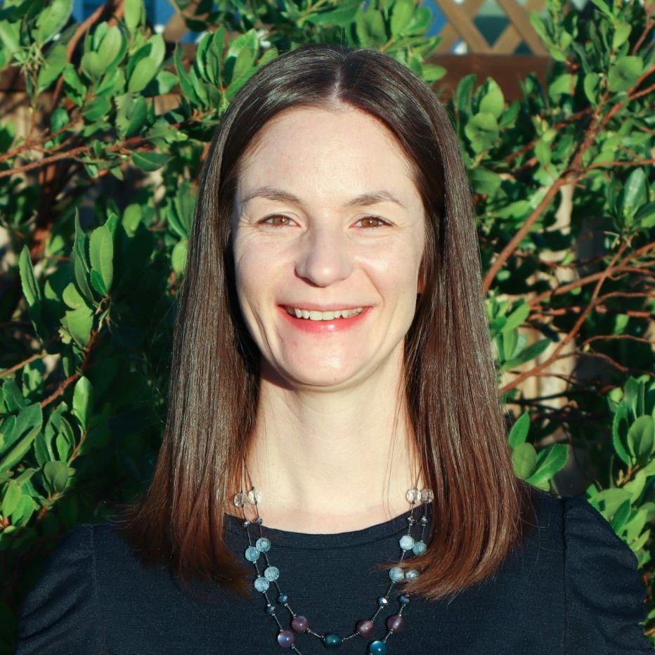Smiling woman with straight brown hair and a beaded necklace, standing outdoors in front of leafy greenery.
