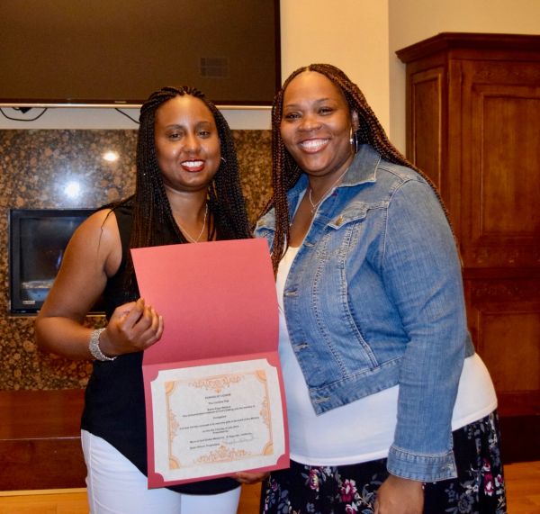 Two women smiling, one holding a certificate inside a red folder.