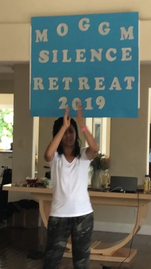 Person clapping under a blue sign reading 'MOGGM SILENCE RETREAT 2019'.