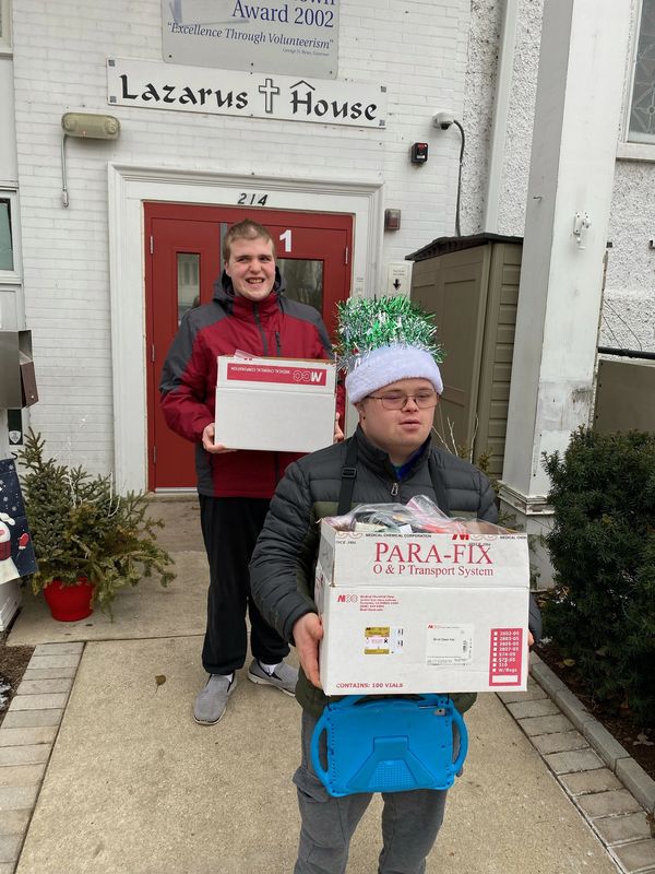 Two men carrying boxes outside Lazarus House, one wearing a festive hat.