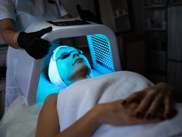 Client under a glowing blue LED light panel during a facial, targeting acne and signs of aging.