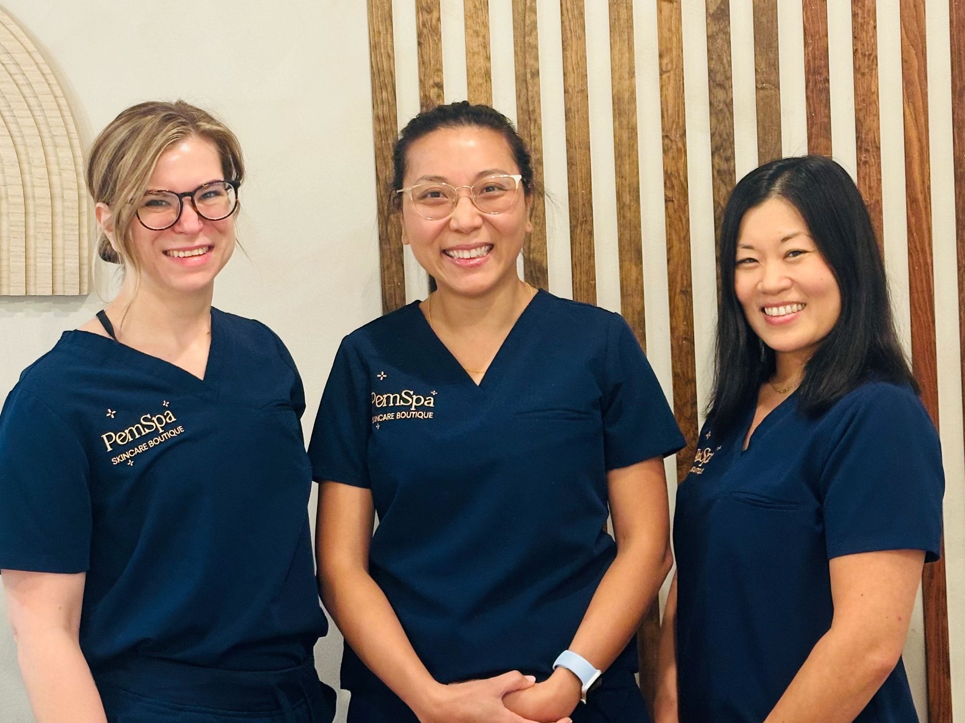 Three PemSpa estheticians smiling in navy uniforms inside the spa with a wood-accented backdrop.