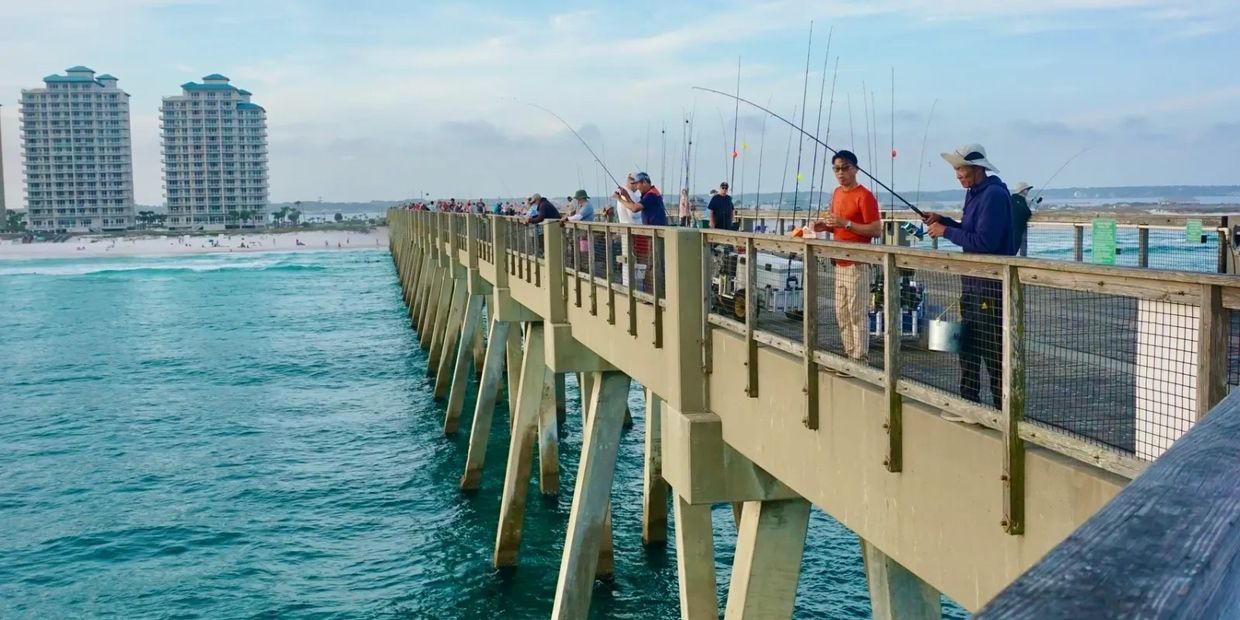 People fishing on a pier by the ocean with buildings in the background.