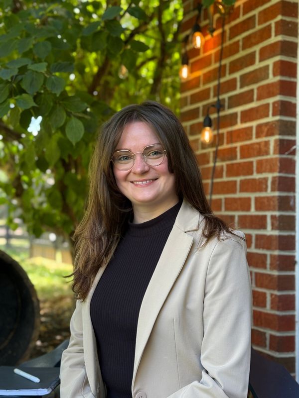 Smiling woman in glasses and beige blazer stands outdoors by a brick wall with hanging lights.