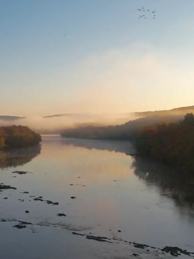 A calm river at sunrise with birds flying overhead and misty hills.