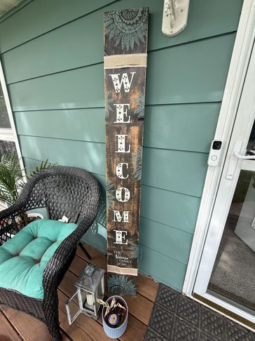 A tall 'Welcome' sign on a porch with a wicker chair and lantern.