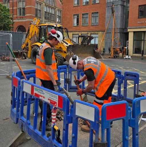 Two construction workers in safety gear working inside a fenced-off street area.
