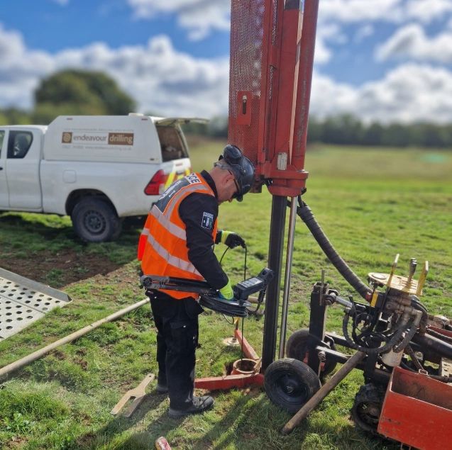 A worker operates a drilling machine on a grassy field wearing safety gear.