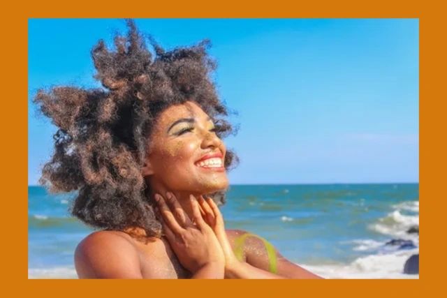 1Brown-skinned woman with natural hair standing at the beach smiling as she looks toward the sky