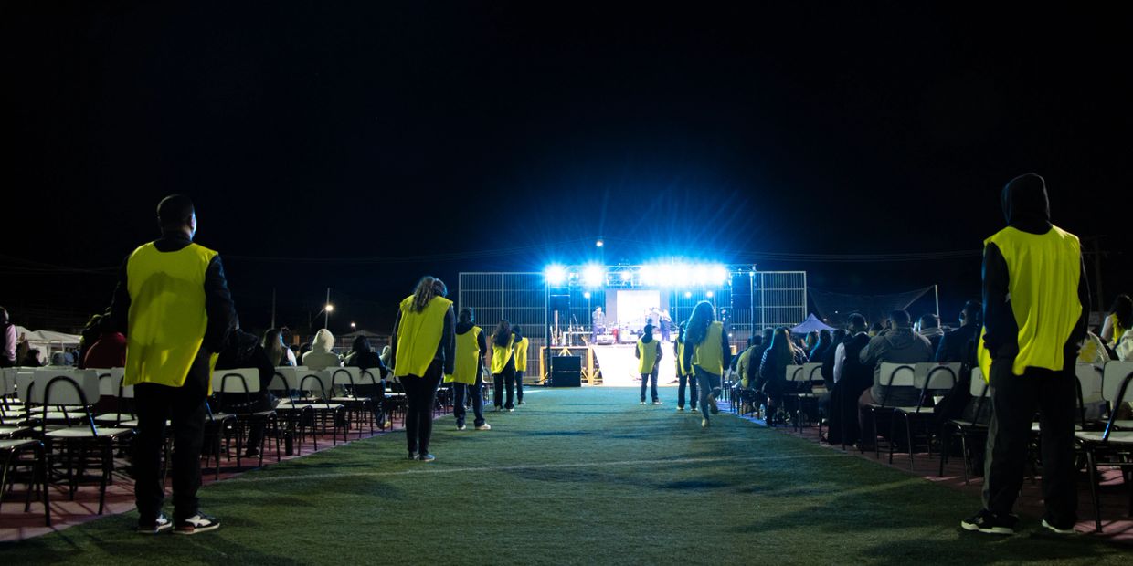 People in yellow vests stand in rows at a nighttime outdoor event with a lit stage.