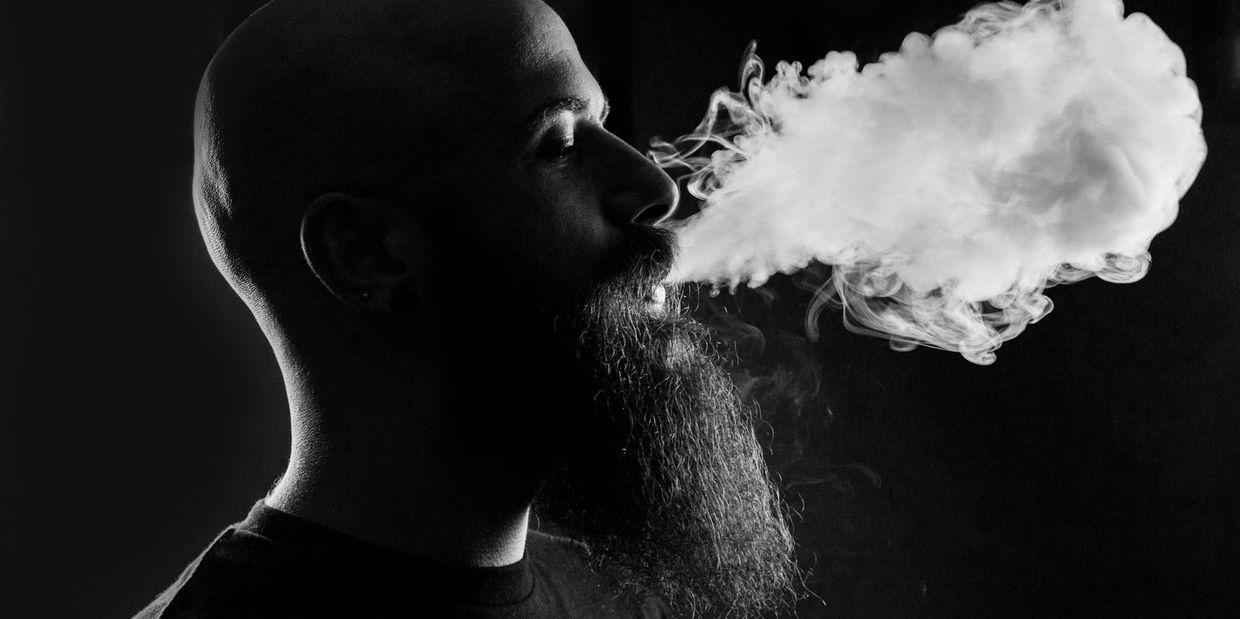 Profile of a bearded man exhaling thick smoke in a dark setting.