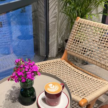A cozy chair next to a table with a coffee cup and a vase of pink flowers.