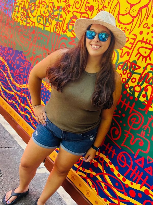 A smiling woman in sunglasses and a hat poses in front of a bright, colorful mural with a lion design.