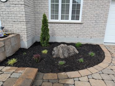 A small garden bed with mulch, plants, and a large rock near a brick house.