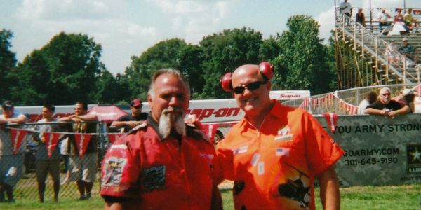 Two men in red shirts posing outdoors at a sunny event.