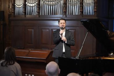 Man speaking into a microphone beside a grand piano in a concert hall.