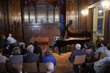 A pianist performs in an elegant room with an audience seated.