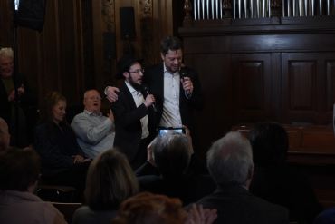 Two men in suits sing with microphones in an audience setting.