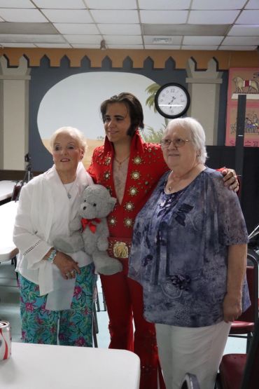 A man dressed as Elvis poses with two elderly women, one holding a teddy bear.