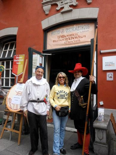 Two men in historical costumes stand with a smiling woman outside a museum entrance.