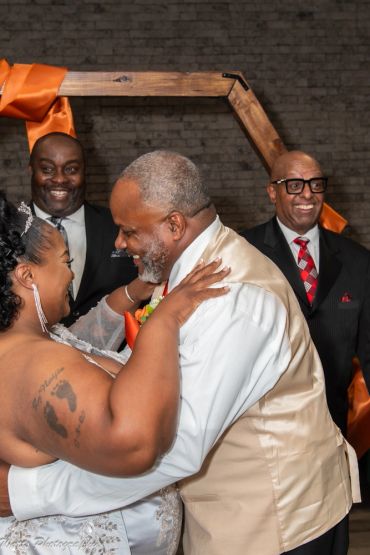 Couple dancing joyfully at wedding with two men smiling behind.