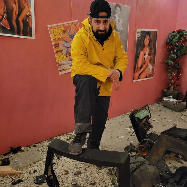 Man in yellow jacket posing indoors with posters on a red wall.