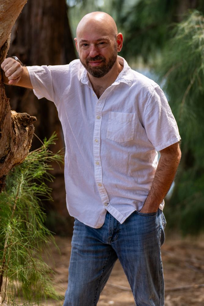 A smiling bald man in a white shirt and jeans leaning on a tree outdoors.