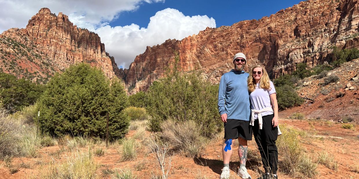 Couple poses smiling in front of red rock cliffs under a partly cloudy sky.