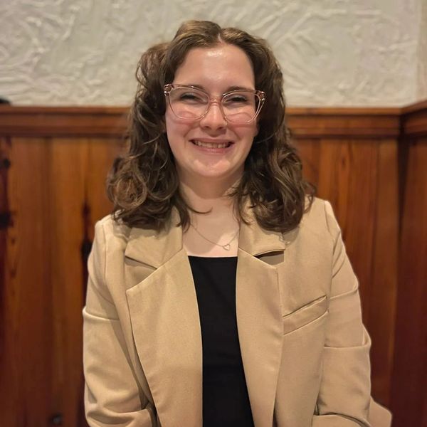 Smiling young woman with glasses and curly hair in a beige blazer.