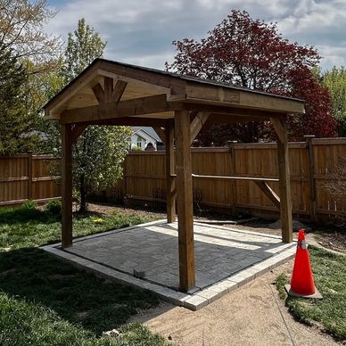 A wooden pergola structure with a stone base in a backyard under a cloudy sky.