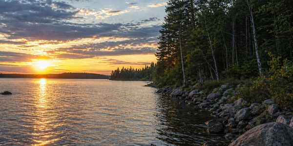 Sunset over a rocky lakeshore with forest trees.