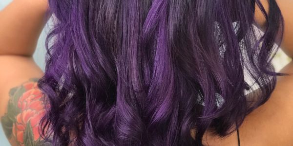 Vibrant purple hair with soft curls and a tattooed arm visible.