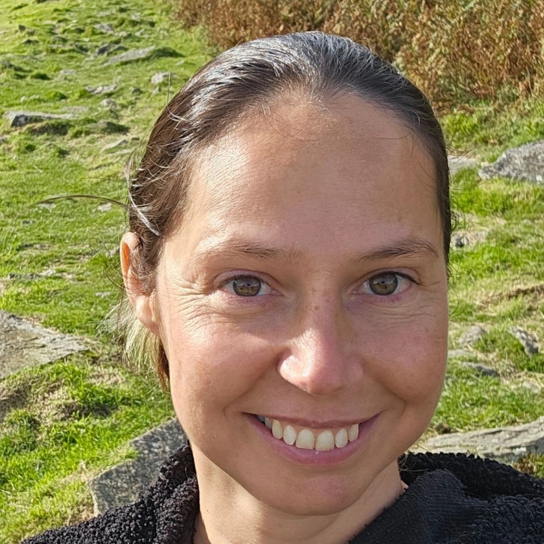Smiling woman in a black hoodie outdoors on a sunny day in a hilly area.
