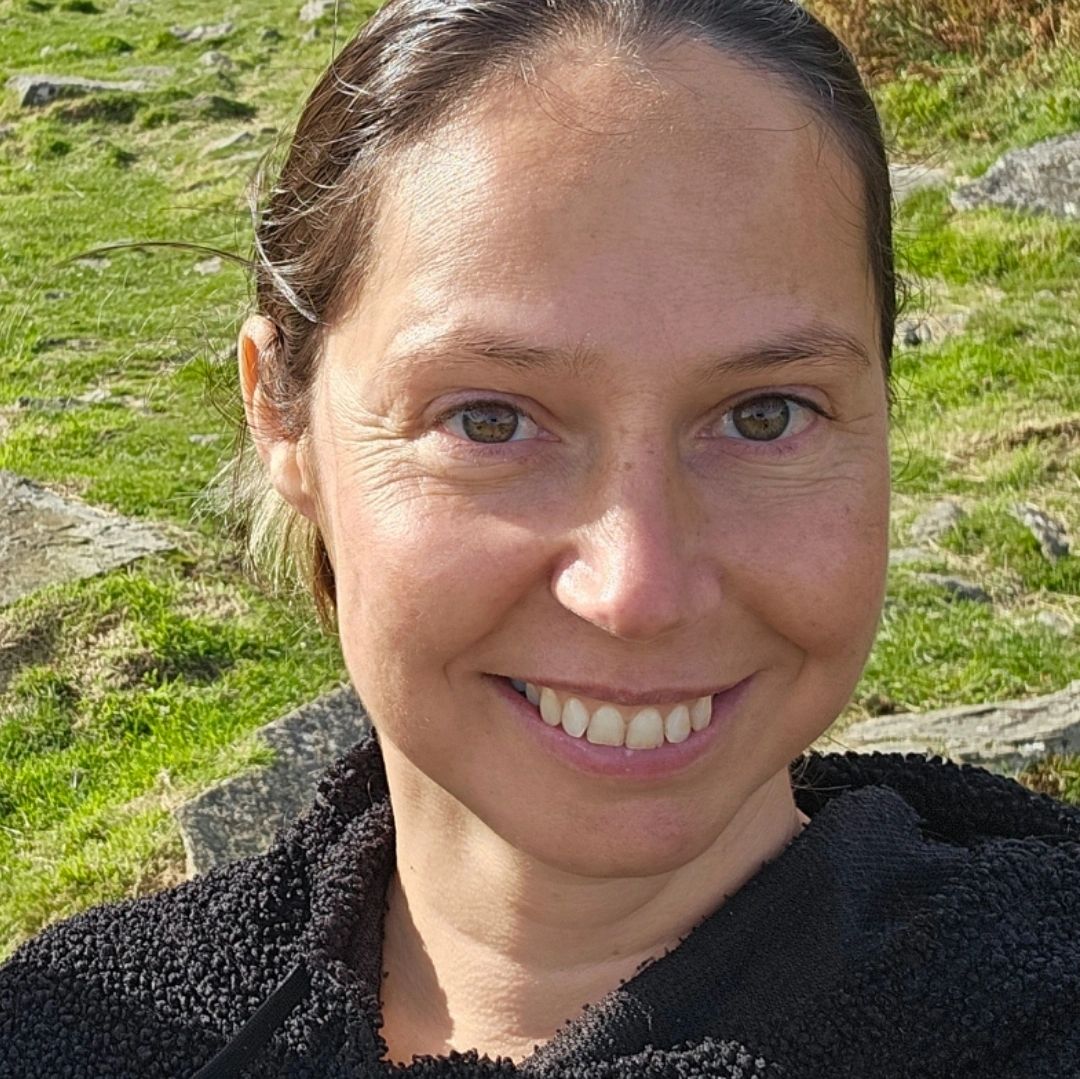 Smiling woman in a black hoodie outdoors on a sunny day in a hilly area.