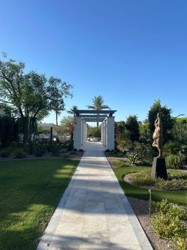 Sunny garden path with white arch and sculpture.